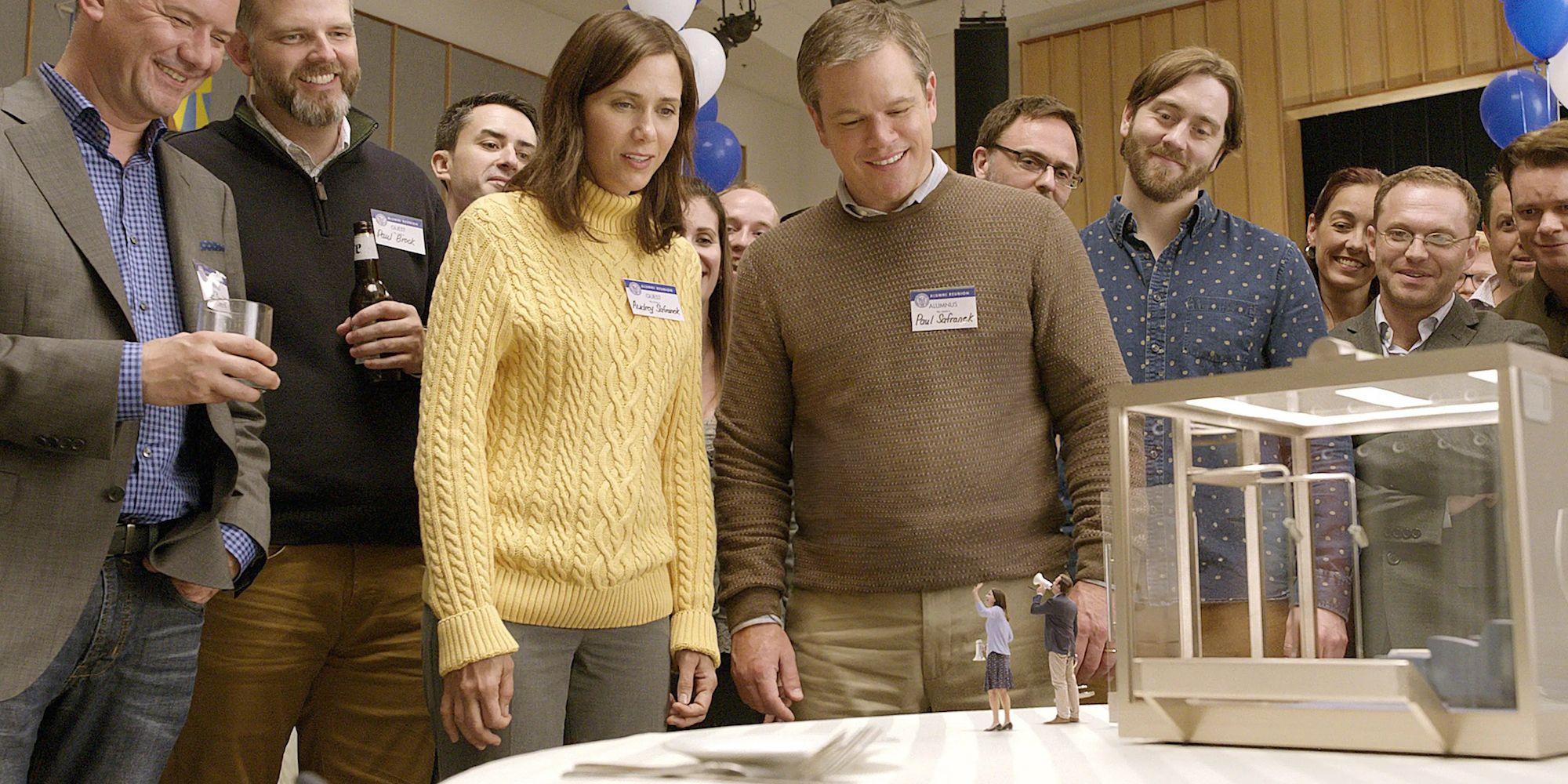 Kristen Wiig and Matt Damon as Audrey and Paul smiling while looking down at two miniature people waving Downsizing